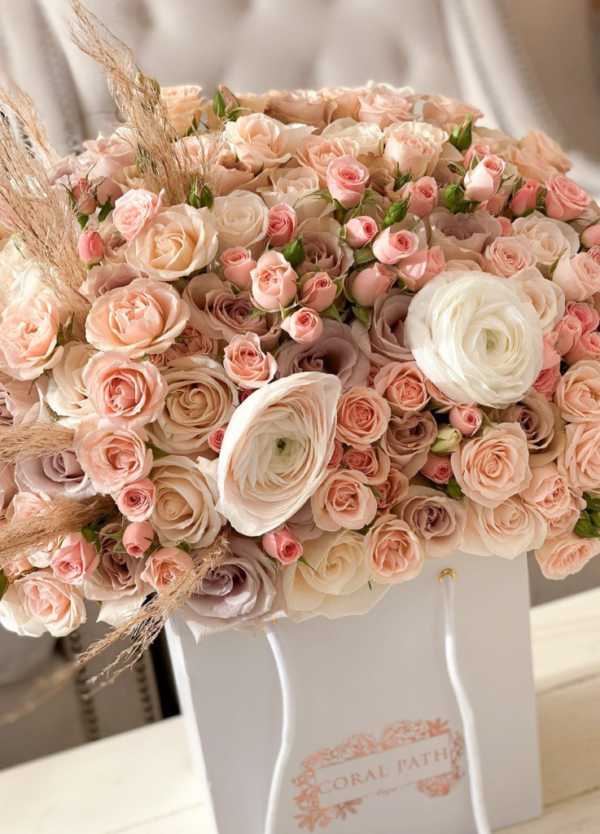 A mix of pastel colored roses and spray roses with pampas grass coming out from the side all in a bag shaped box flower arrangement