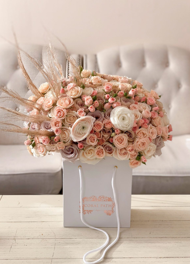 A mix of pastel colored roses and spray roses with pampas grass coming out from the side all in a bag shaped box flower arrangement