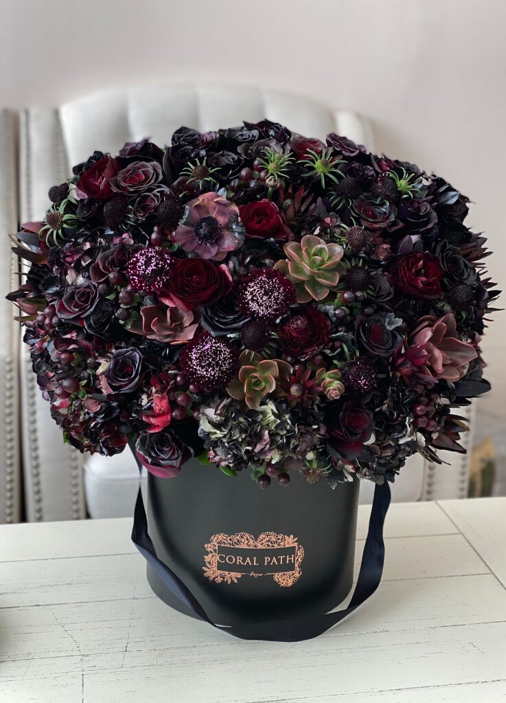 Dark burgundy and black flower mix arranged in a hat box with a logo.