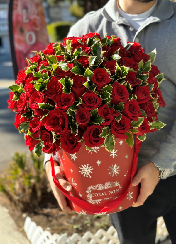 Image description: Person holding flowers. Christmas box with red roses, holly greenery, and silver snowflakes, bringing warmth and festivity to the holidays."