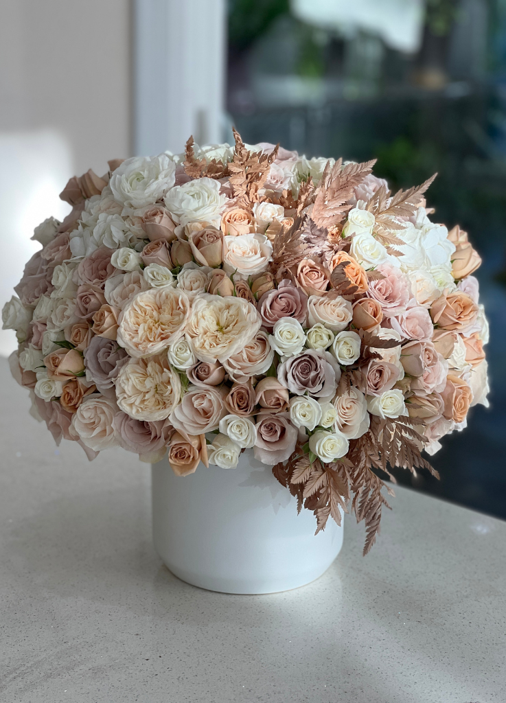 Image description: Stylish flower arrangement in a ceramic vase featuring roses, garden roses, and dried leaves in neutral tones.