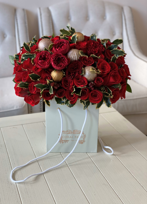 Image description: Festive flower arrangement in a bag-shaped box with red roses, Christmas ornaments, and holly leaves for holiday celebrations.