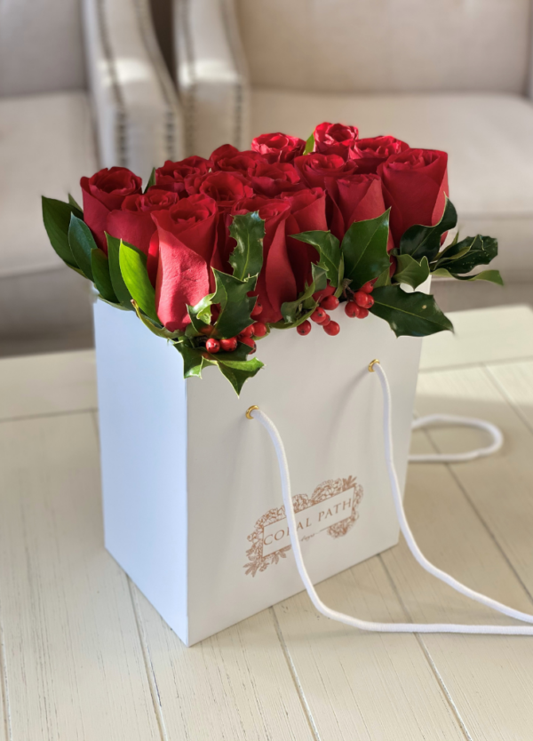 Image description: Festive bag-shaped flower arrangement with red roses and holly greenery, perfect for holiday celebrations and joyous occasions.