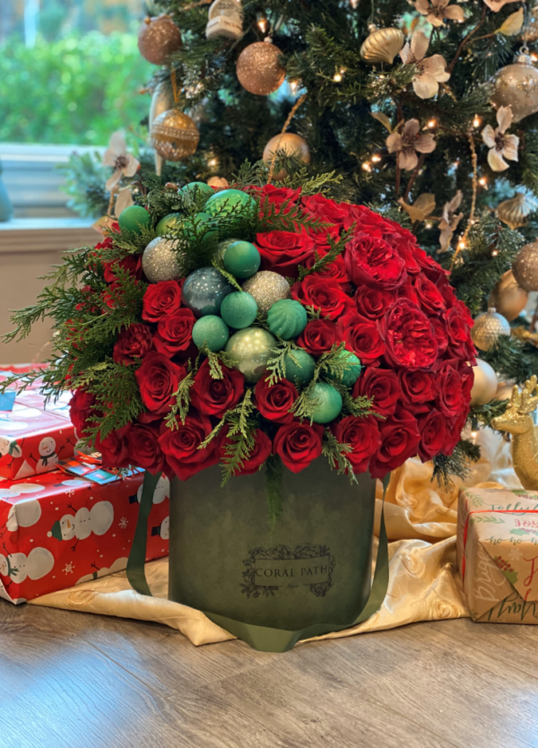 "Image description: Circular hat box arrangement in Victoria Green with red roses, Christmas ornaments, and fern leaves for festive elegance.