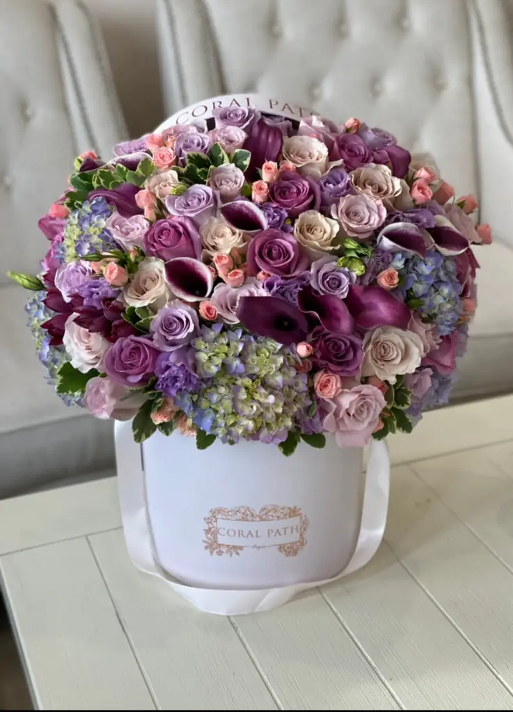 A set of purple and lavender flowers such as calla lilies, hydrangeas and roses arranged in a hat box.
