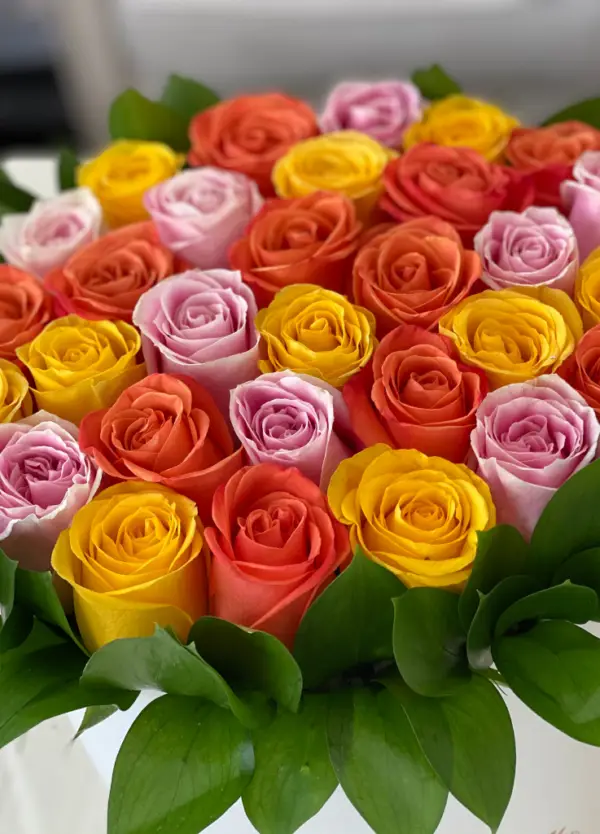 Orange, yellow and pink roses arranged in a hat box with green leaves around it.