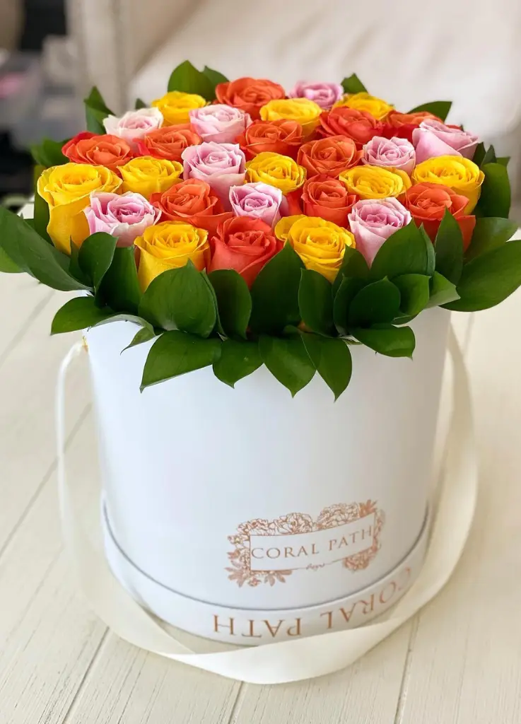 Orange, yellow and pink roses arranged in a hat box with green leaves around it.