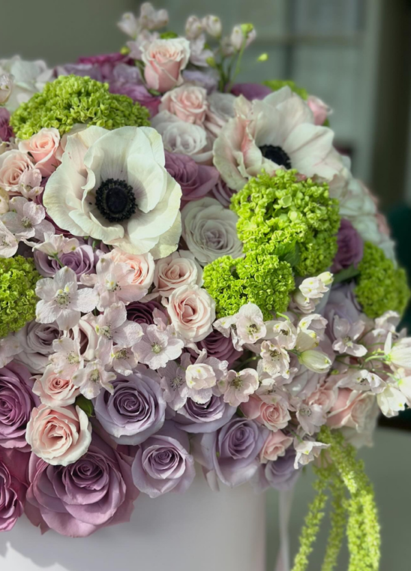 Lavender roses, amaranthus, anemones, seasonal blooms, in a French hat box—a garden's charm captured in 'Du Jardin' arrangement.