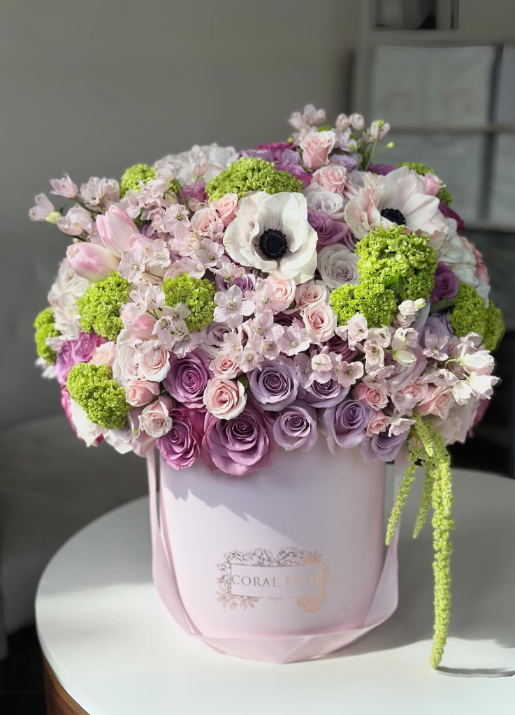 Lavender roses, amaranthus, anemones, seasonal blooms, in a French hat box—a garden's charm captured in 'Du Jardin' arrangement.