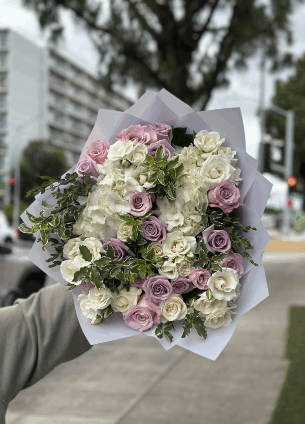 Lavender and ivory rose bouquet with hydrangea and greenery, wrapped in white paper and held outdoors on a city street.