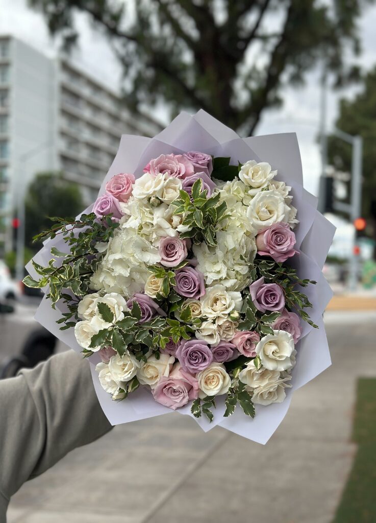 Lavender and ivory rose bouquet with hydrangea and greenery, wrapped in white paper and held outdoors on a city street.