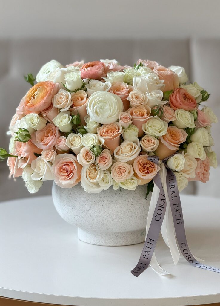 Lush peach and cream garden rose arrangement with ranunculus and spray roses in a rounded ceramic vase, finished with Coral Path ribbon.