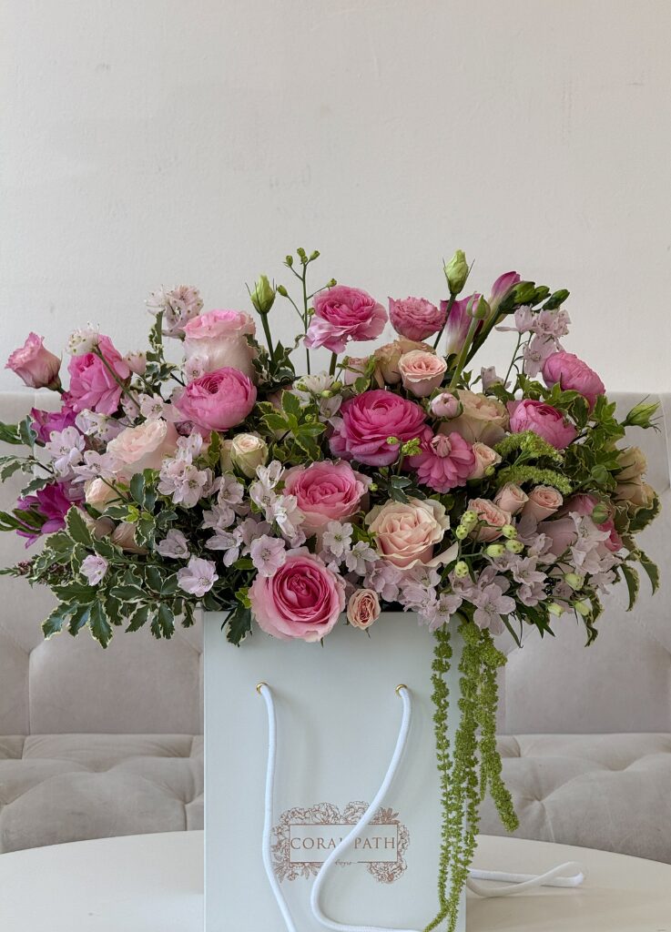 Pink garden rose and ranunculus arrangement in a white Coral Path hat box with cascading greenery and soft seasonal blooms.
